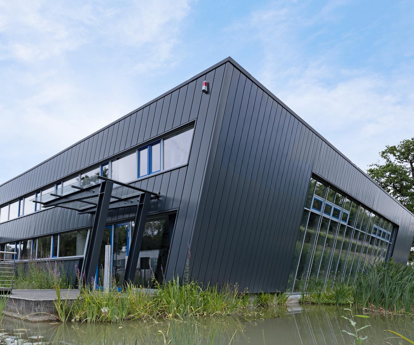 Moderne, schwarze Gebäudeansicht mit großen Fenstern, umgeben von grasbewachsenen Uferbereichen und Wasserpflanzen.