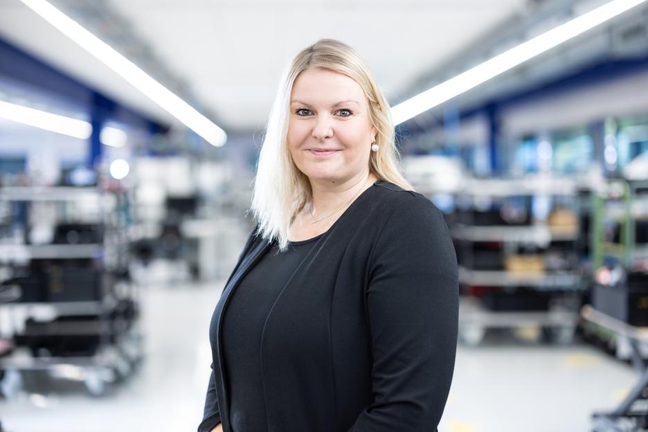 Porträt einer lächelnden Frau mit blonden Haaren in einem modernen, industriellen Umfeld mit Regalen im Hintergrund.