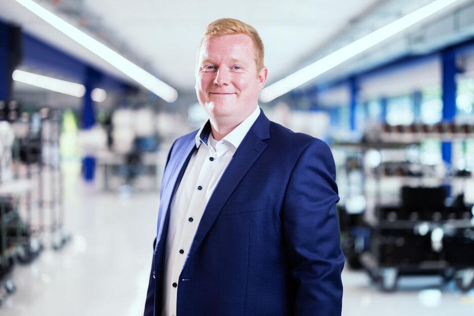 Mann in Anzug lächelt in einem modernen, hellen Raum mit Regalen im Hintergrund.