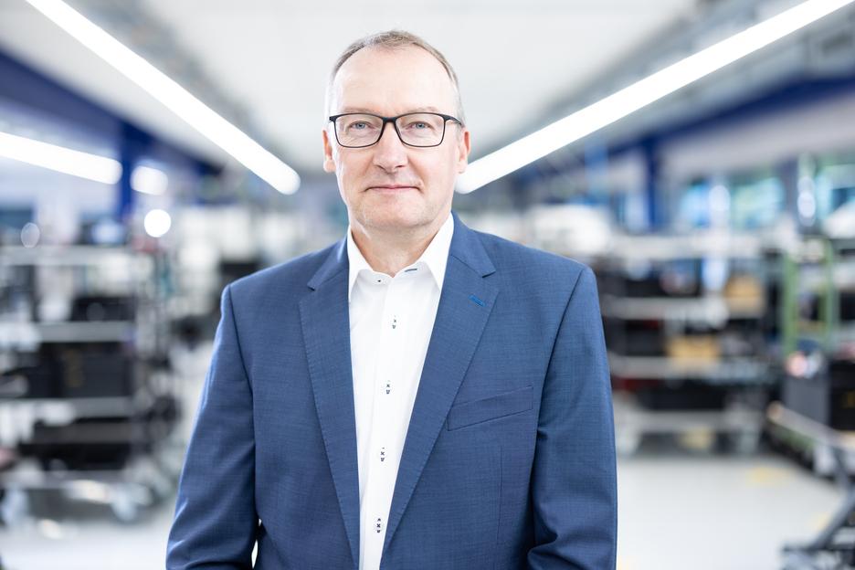 Porträt eines Mannes in einem blauen Anzug vor einem industriellen Hintergrund mit Regalen und gedämpften Lichtern.