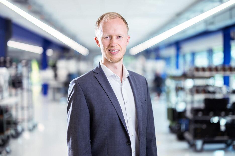 Porträt eines Mannes in einem grauen Anzug, vor einem modernen, hellen Hintergrund mit Regalen und Arbeitsbereichen.