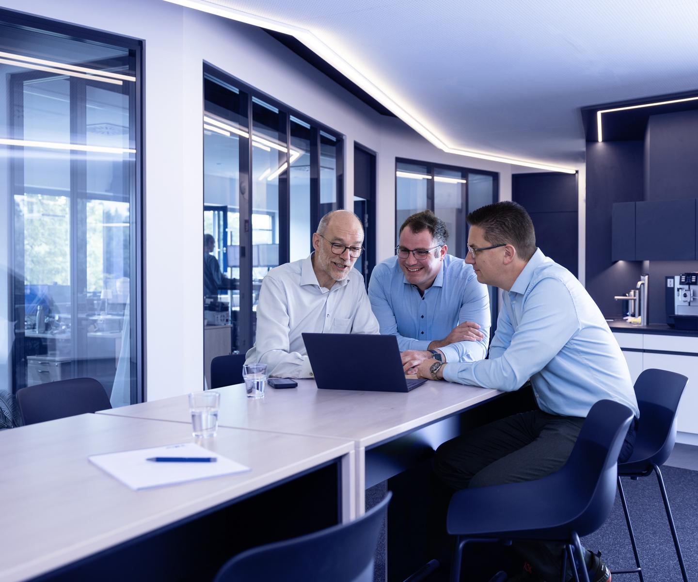 Drei Männer in Hemden sitzen an einem Tisch und betrachten gemeinsam einen Laptop im modernen Büro.