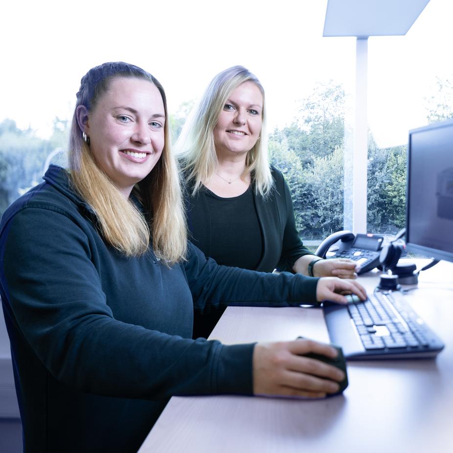 Zwei Frauen sitzen an einem Schreibtisch, arbeiten am Computer und lächeln in die Kamera. Hintergrund mit Bäumen.