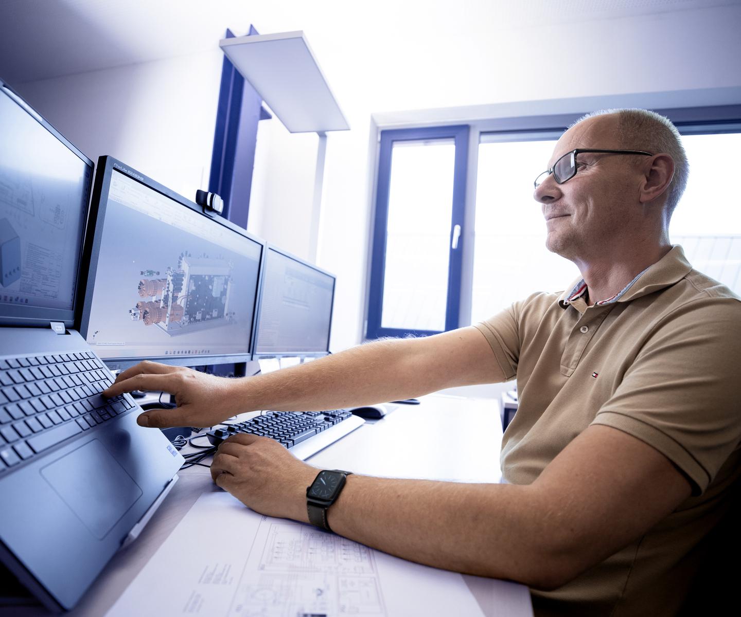 Mann mit Brille arbeitet an mehreren Monitoren, die technische Zeichnungen und Modelle zeigen.
