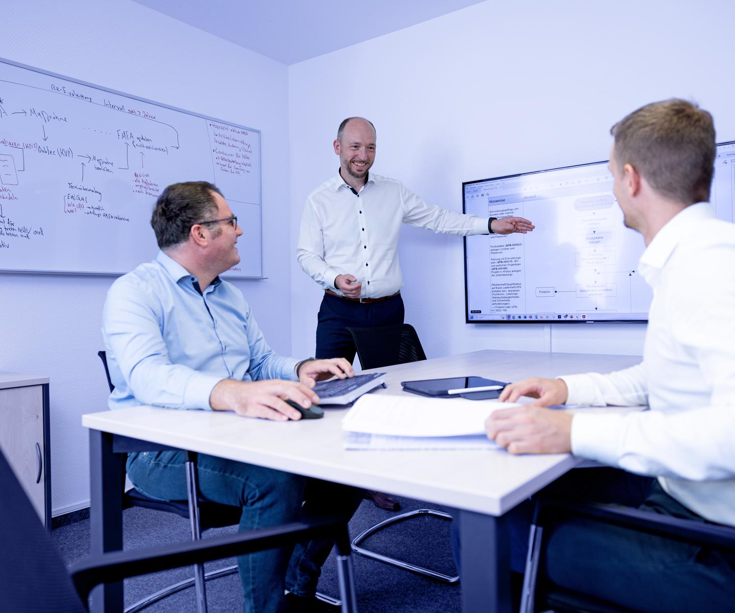 Drei Personen in einem Meetingraum diskutieren, während einer an einem Bildschirm eine Präsentation zeigt.