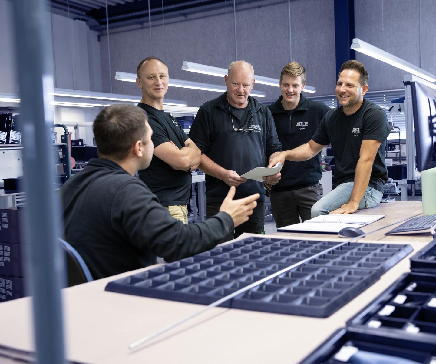 Gruppe von fünf Männern in Arbeitskleidung diskutiert um einen Tisch mit Dokumenten und Werkzeugen in einer Werkstatt.