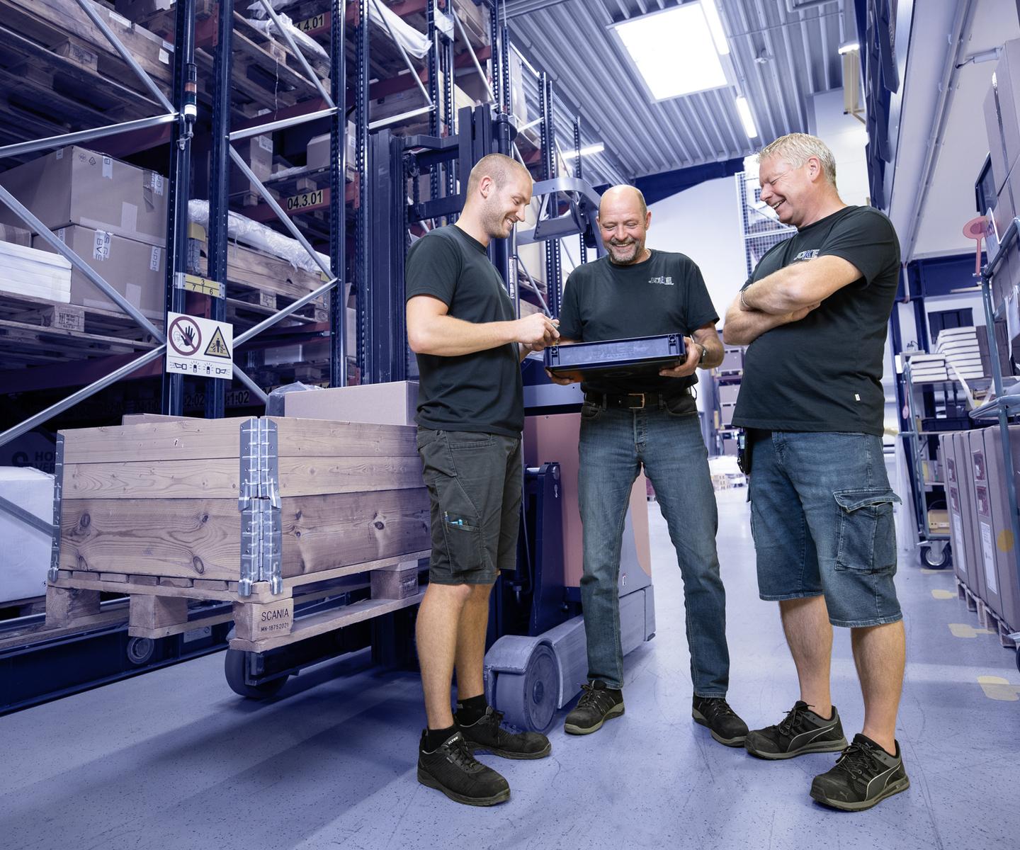 Drei Männer in Arbeitskleidung schauen auf einen Laptop in einem Lager mit Regalen voller Kisten und Paletten.