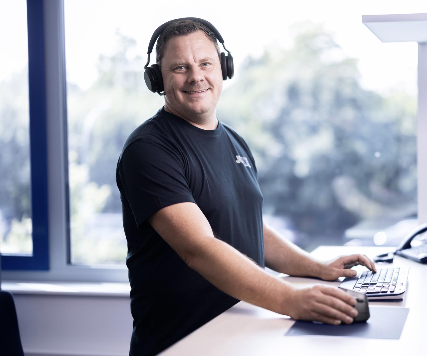 Mann mit Kopfhörern arbeitet an einem Schreibtisch mit Computern und Telefon in einem hellen Büro.