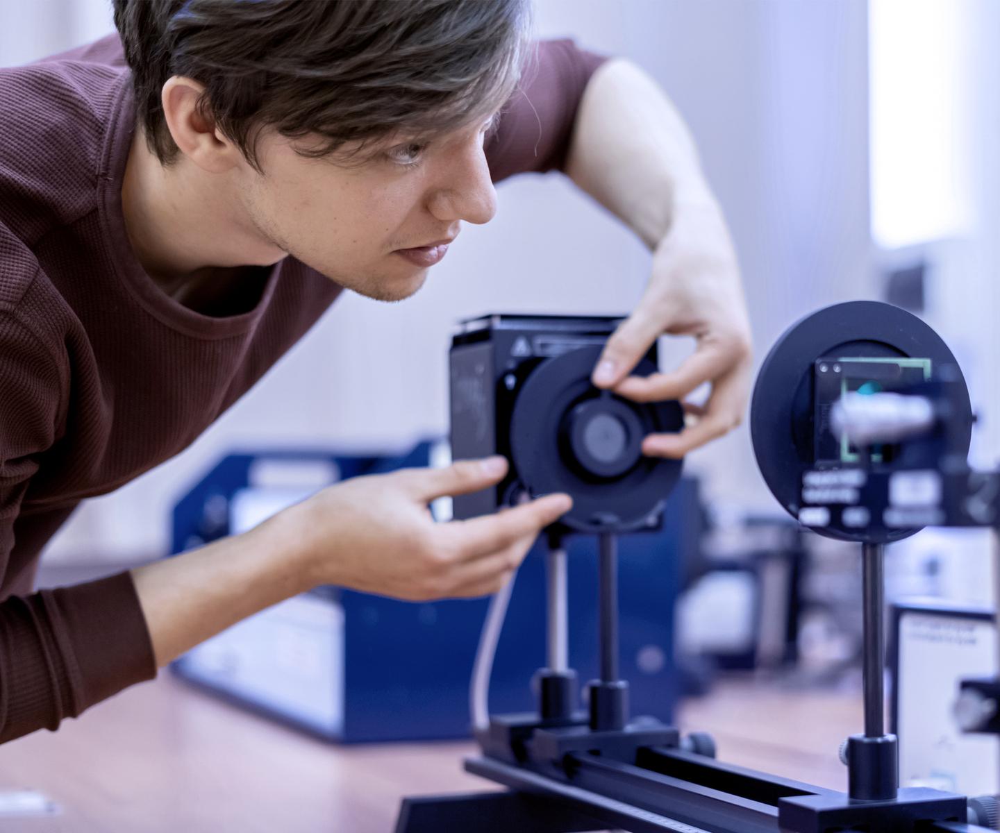Junger Mann justiert ein optisches Gerät auf einem Tisch in einem Laborumfeld.