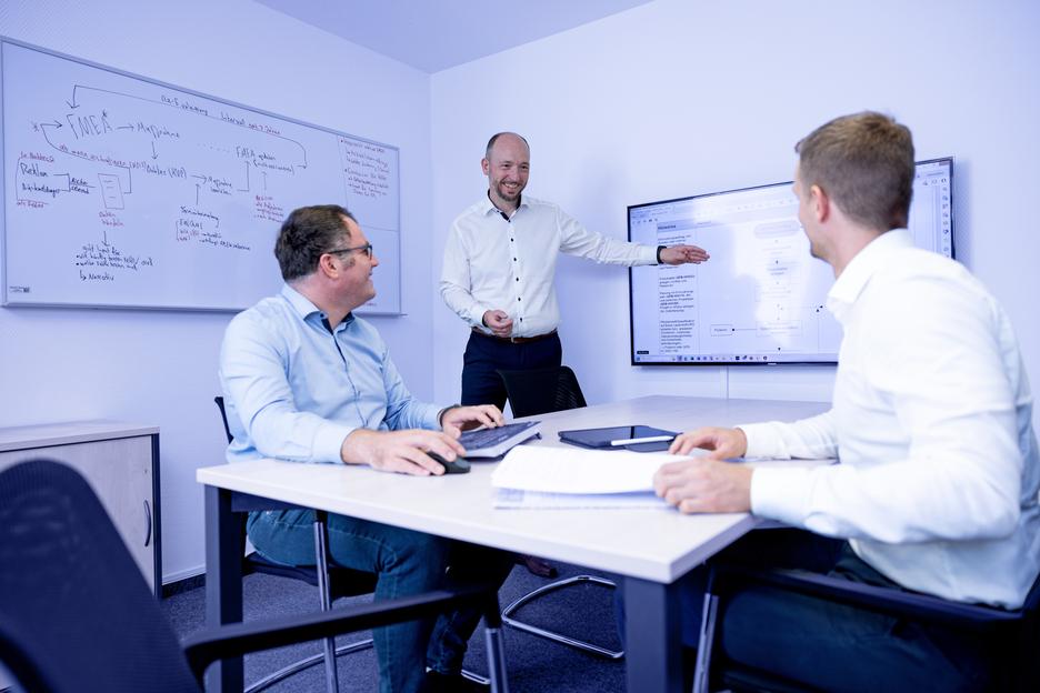 Drei Personen in einem Meetingraum diskutieren, während einer an einem Bildschirm eine Präsentation zeigt.