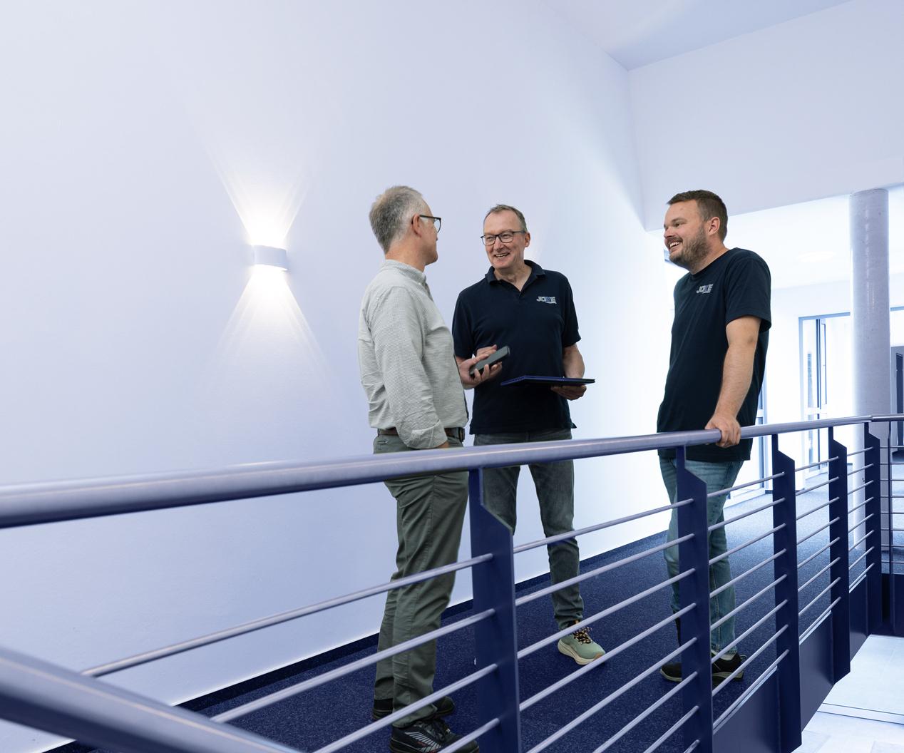 Drei Männer stehen auf einer Treppe und unterhalten sich, ein Tablet in der Hand, im modernen, hellen Raum.
