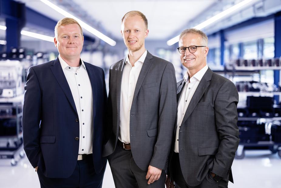 Drei Männer in Anzügen stehen in einem modernen Büro mit technischen Geräten im Hintergrund.
