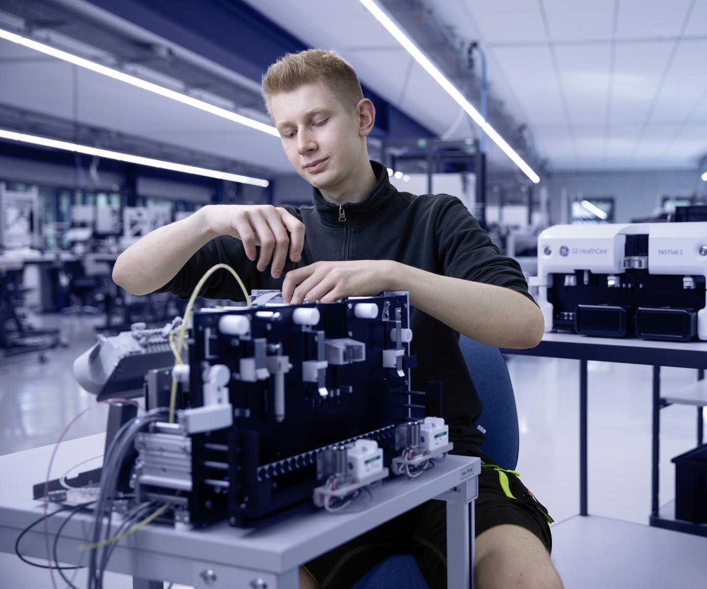 Junger Mann arbeitet an einem technischen Gerät in einem modernen Labor mit medizinischen Geräten im Hintergrund.