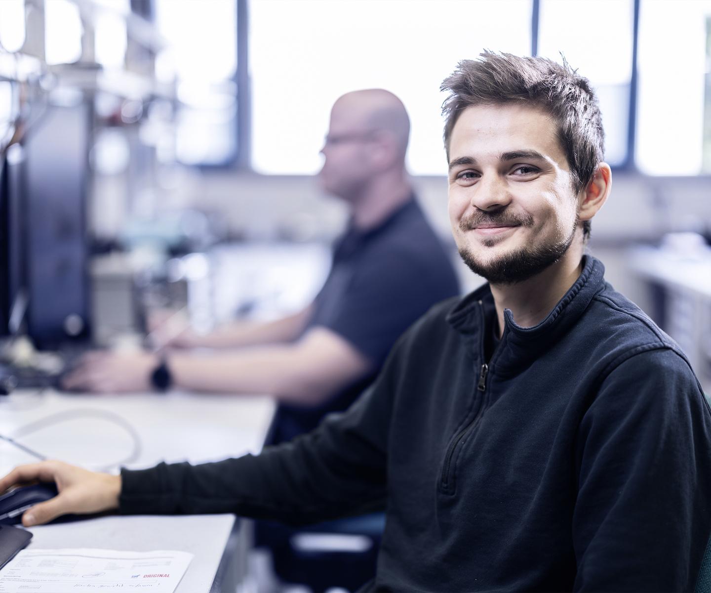 Lächelnder junger Mann sitzt an einem Schreibtisch, vor ihm ein Computer, im Hintergrund ein weiterer Kollege.