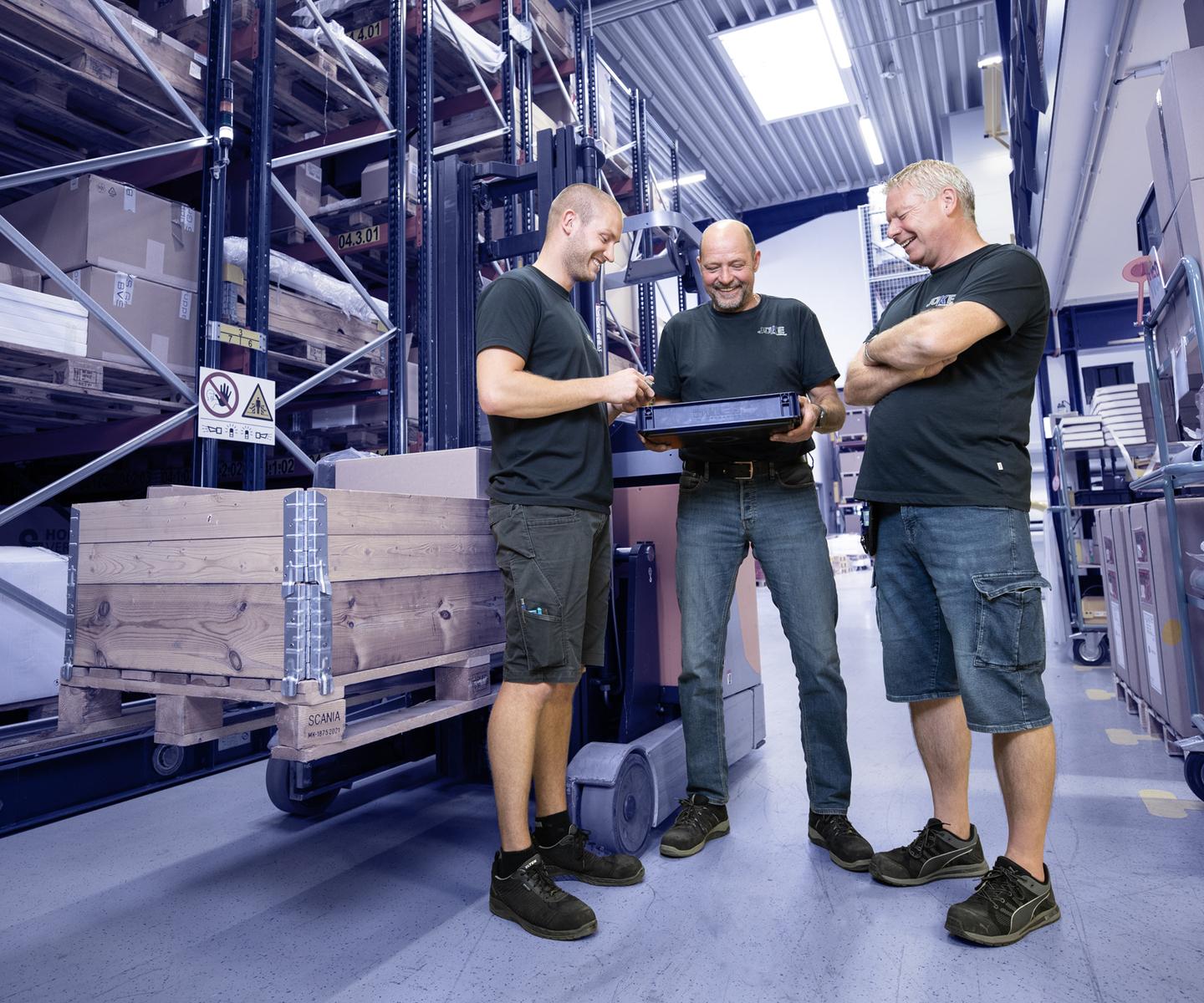 Drei Männer in Arbeitskleidung schauen auf einen Laptop in einem Lager mit Regalen voller Kisten und Paletten.