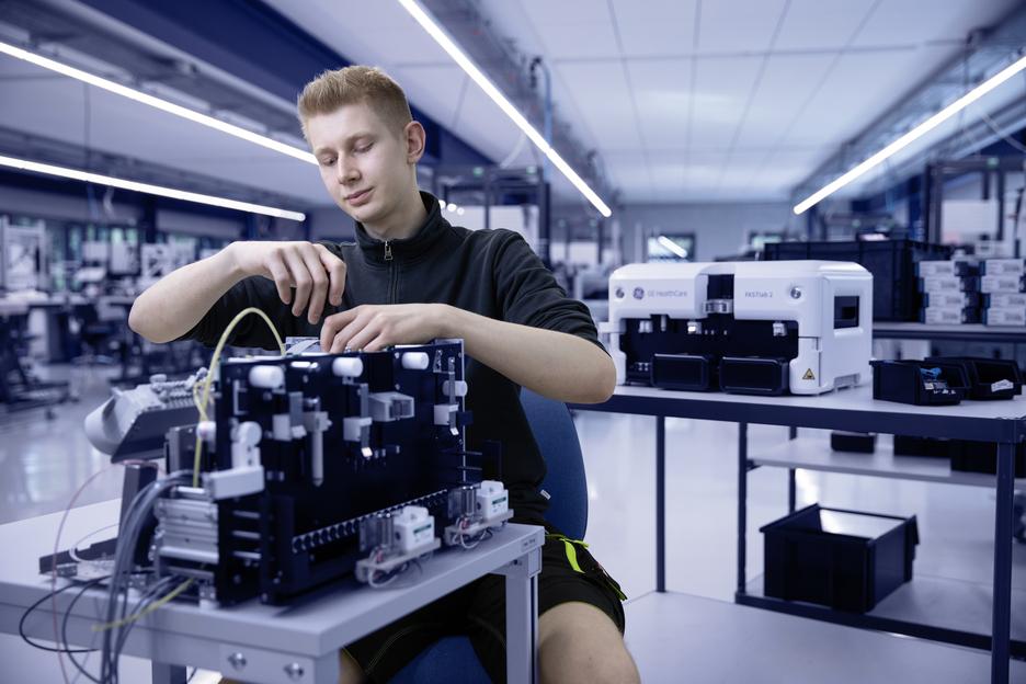 Junger Mann arbeitet an einem technischen Gerät in einem modernen Labor mit medizinischen Geräten im Hintergrund.