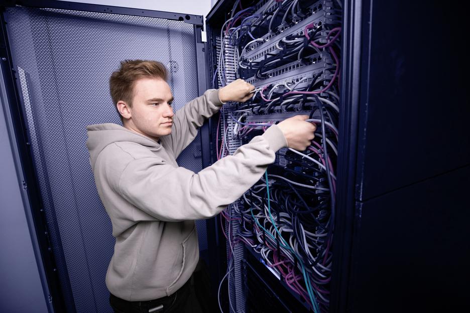 Junger Mann in grauem Hoodie verkabelt ein Server-Rack mit bunten Kabeln.