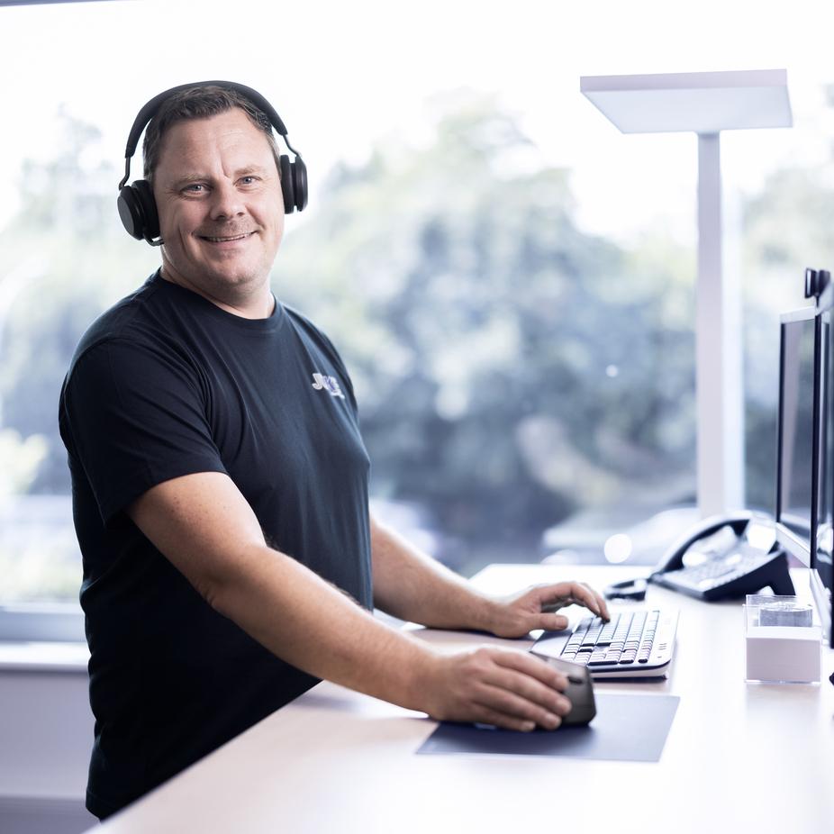 Mann mit Kopfhörern arbeitet an einem Schreibtisch mit Computern und Telefon in einem hellen Büro.