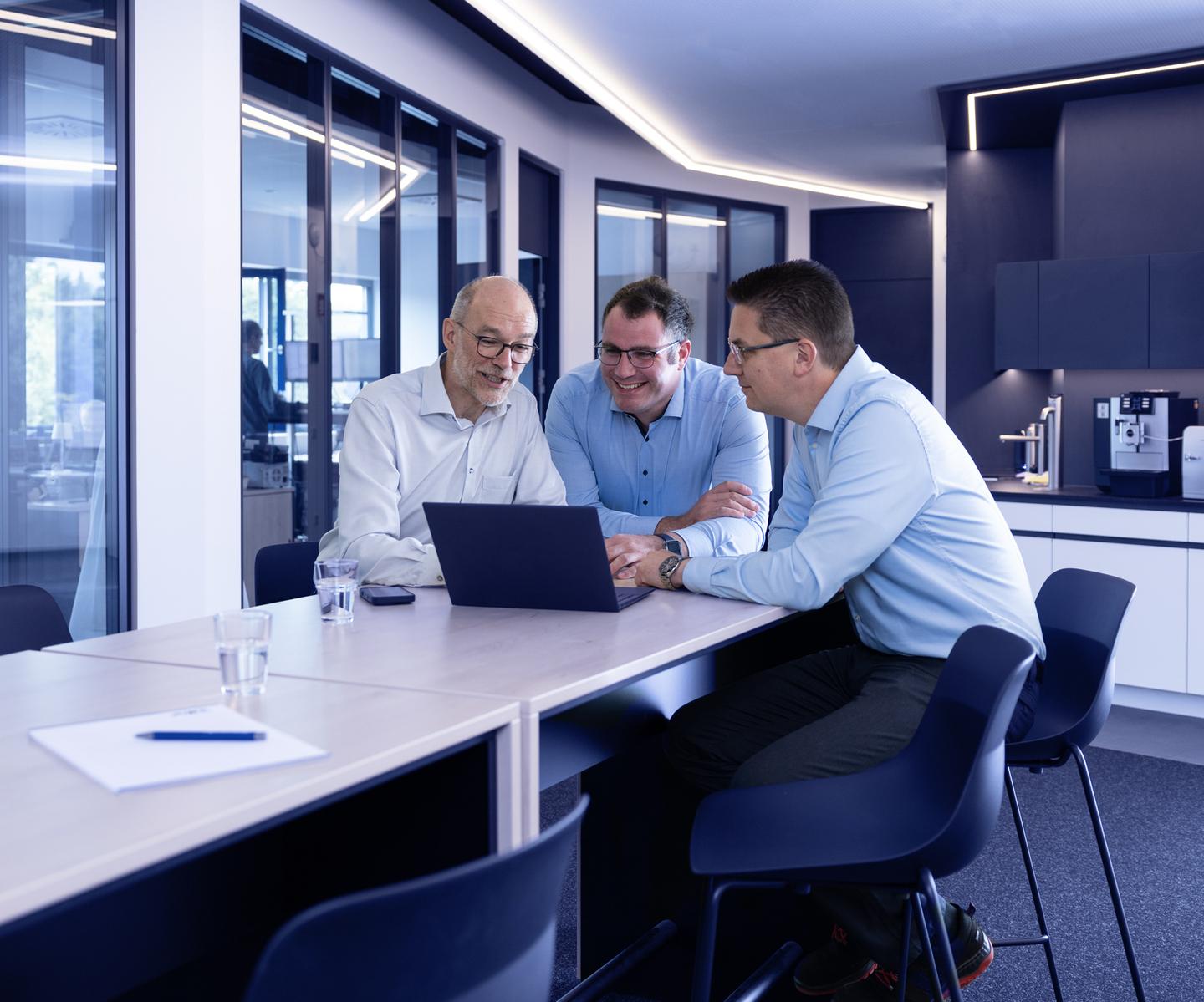 Drei Männer in Hemden sitzen an einem Tisch und betrachten gemeinsam einen Laptop im modernen Büro.