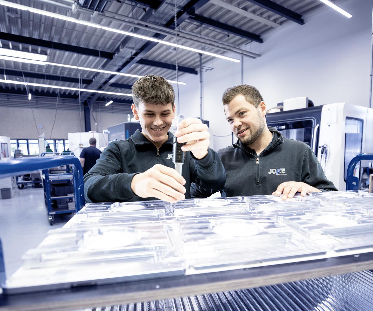 Zwei Männer überprüfen gemeinsam transparente Teile auf einem Werkstattwagen in einer modernen Fertigungsumgebung.