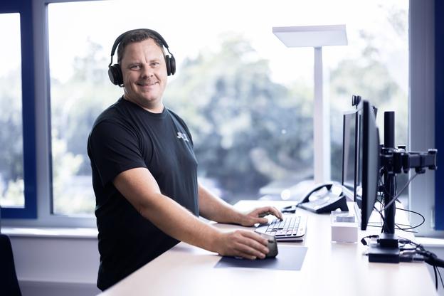 Mann mit Kopfhörern arbeitet an einem Schreibtisch mit Computern und Telefon in einem hellen Büro.
