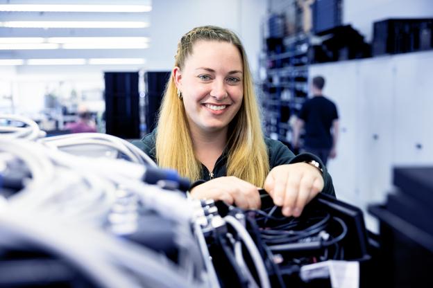 Frau mit langen, blonden Haaren arbeitet lächelnd an einer Verkabelung in einem modernen technischen Raum.