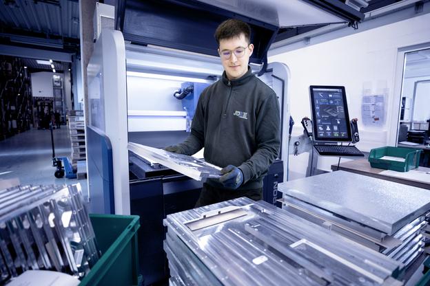 Junger Mann in Arbeitskleidung hält bearbeitete Metallplatten vor einer Maschine in einer Produktionshalle.