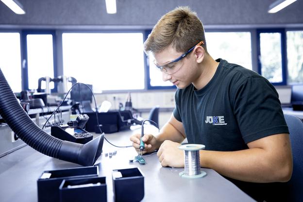 Junger Mann mit Schutzbrille lötet an einer Leiterplatte in einer Werkstatt, mit Kabeln und Werkzeugen auf dem Tisch.