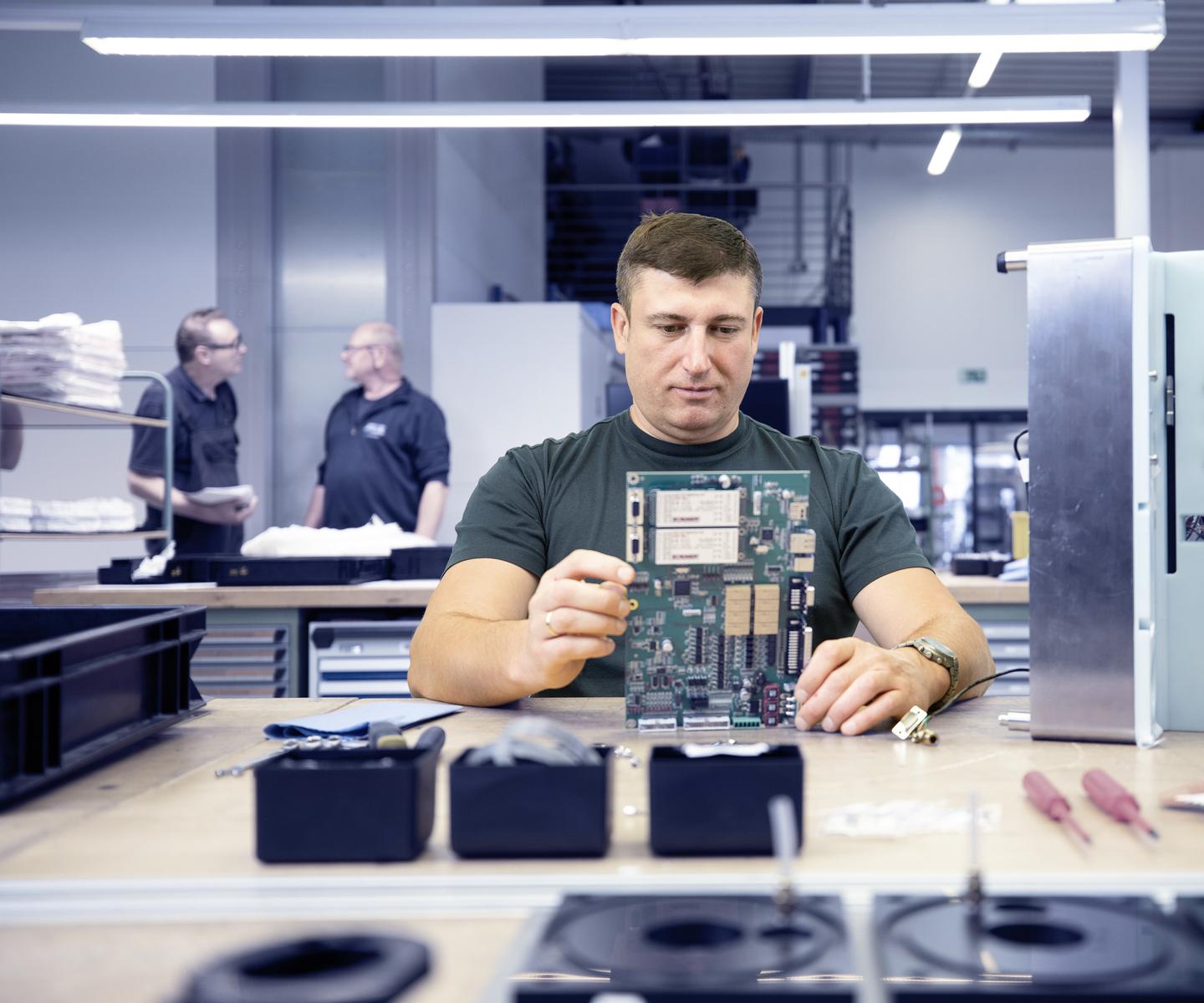 Mann in T-Shirt betrachtet eine Platine an einem Tisch, während zwei Kollegen im Hintergrund miteinander sprechen.