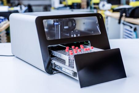 Moderne Labormaschine mit geöffnetem Fach für Glasfläschchen auf einem Tisch in einem technischen Umfeld.